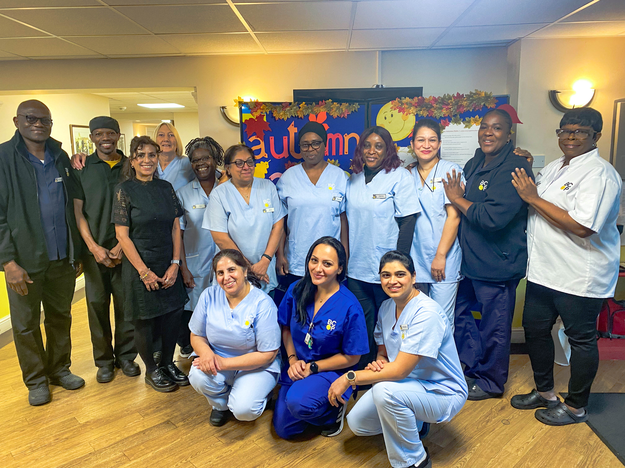 Members of Robert Harvey House Staff team. Team members are dressed differently based on their role, with care workers in light blue, nursing staff in royal blue and maintenance staff in Navy blue. Catering staff have on white chef overalls Office and administrative staff are dressed in business casual. Most members arae standing with three members of staff squatting at the front.